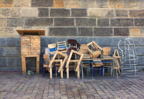 A collection of discarded furniture and wooden items resting against a weathered brick wall outdoors. The assortment includes various wooden chairs, some with distinctive painted finishes, stacked and leaning in different directions. There are also disassembled wooden tables with legs, and a small wooden cabinet or storage unit with a slatted door positioned on the left side. To the far right, a metallic folding chair and a metal plant stand with multiple circular tiers are visible. The ground is paved with irregular stone or cobblestone blocks, and the environment appears to be an outside area, possibly in Manor Park where garden clearance services are often needed. The lighting is natural, suggesting daytime, and the overall scene depicts waste or rubbish that would typically be handled by professional clearance or rubbish removal services, such as those offered by Garden Clearance Manor Park.