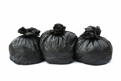 Three large black rubbish bags made of thick plastic, tightly tied at the top, arranged in a row on a plain white background. The bags appear to contain garden or household waste, indicated by their slightly bulging shapes and crinkled surface textures. The middle bag is positioned slightly higher than the others, creating a small gap between them. The bags are clean and well-sealed, with no visible tears or leaks, emphasizing their readiness for disposal. The simple, neutral setting highlights the bags as the primary focus, reflecting typical waste collection or garden clearance scenarios in the Manor Park area. This image aligns with the type of rubbish removal services provided by Garden Clearance Manor Park, used to visually communicate their capacity to handle garden and household waste removal in the local community.