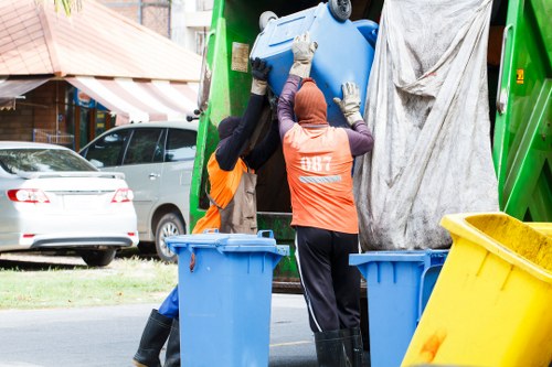 Two waste management workers wearing high-visibility orange vests and gloves are lifting a large blue recycling bin into the back of a green rubbish collection vehicle parked on a residential street. One worker is positioned on the left, with their back partially visible, while the other is more centrally located, facing away but with their head turned slightly to the right. The vehicle's rear door is open, revealing a grey tarp hanging inside. In the foreground, three additional bins are visible, including a yellow bin on the right and two blue bins to the left. The scene is set on a roadway with parked cars, a grassy verge, and residential houses with front gardens and striped awnings in the background. The environment appears calm and typical of a suburban area, with natural daylight illuminating the scene, emphasizing the textures of the bins, the workers' clothing, and the vehicle. The image captures the process of rubbish collection, associated with services provided by Garden Clearance Manor Park in the local area.