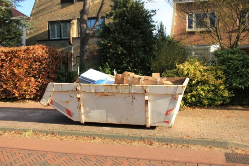 A large white metal skip container placed on a paved driveway in a residential area, filled with broken cardboard boxes and construction debris, with visible signs of rust and wear on its surface. The skip is positioned adjacent to a brick-paved footpath, and behind it, there are trees, shrubs, and detached houses with pitched roofs, typical of a suburban neighbourhood. The surrounding environment suggests a garden clearance or renovation waste collection, with natural daylight illuminating the scene. This image illustrates typical rubbish removal activities undertaken by Garden Clearance Manor Park, serving local residents in the area near Postcode inferred from the context, possibly within the Manor Park vicinity.