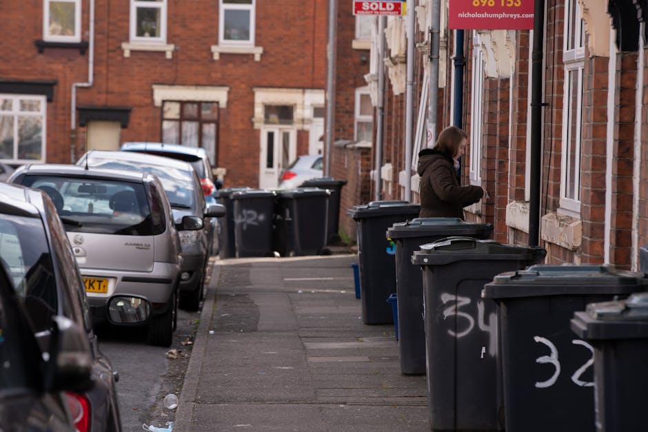 A large collection of overflowing rubbish and waste materials is piled on a paved area in front of a parking lot, consisting of cardboard boxes, plastic bags, crumpled paper, and various discarded pac